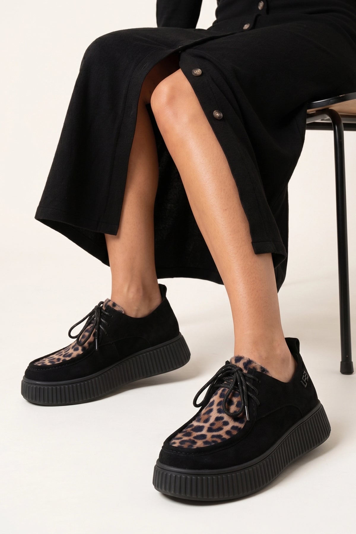 Derbies Peline noir/taupe léo portées avec une jupe noire fendue, assise sur une chaise noire.