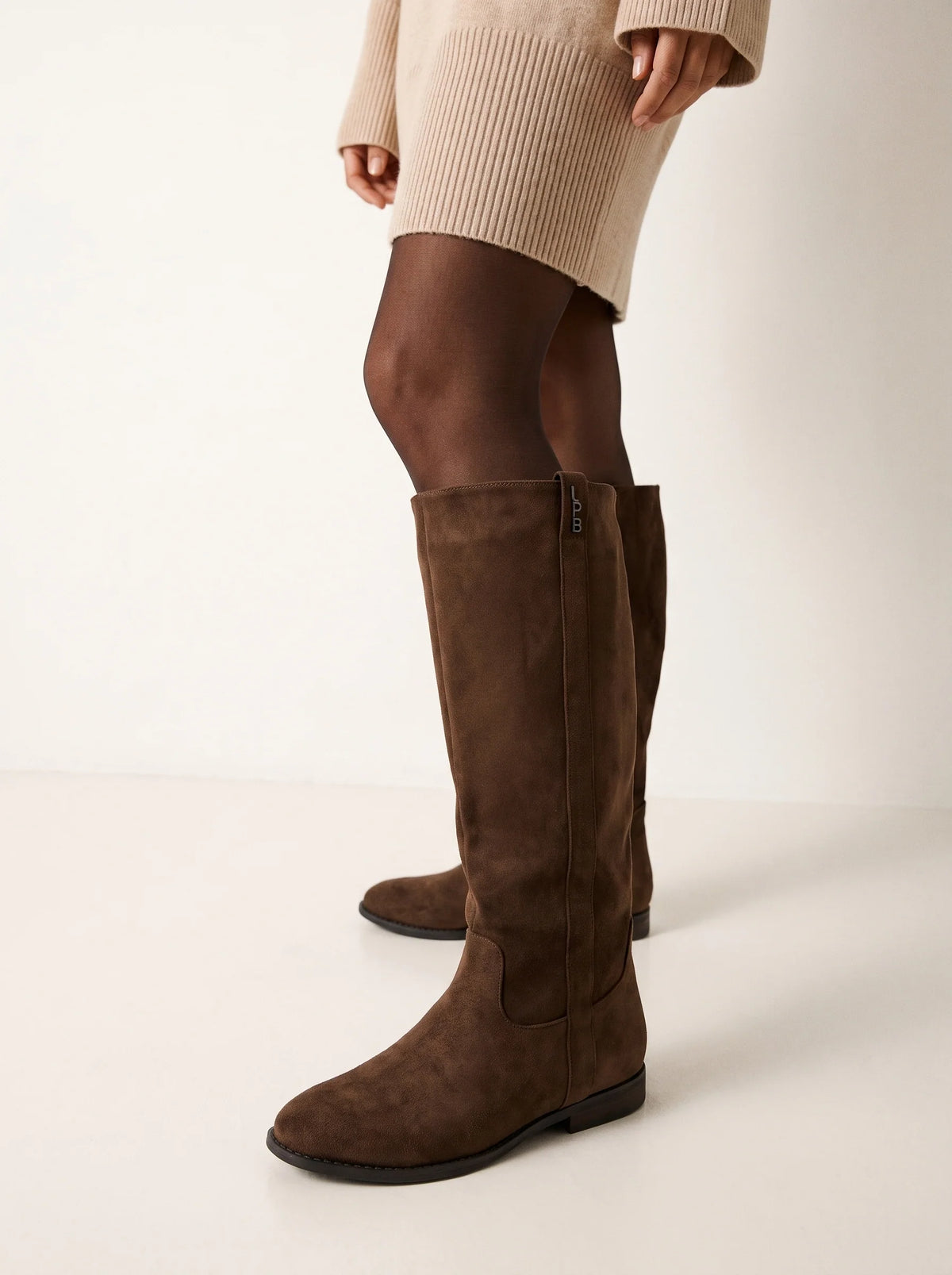 Bottes Pauline choco portées avec une robe beige, collant noir transparent, tige haute en suédine.